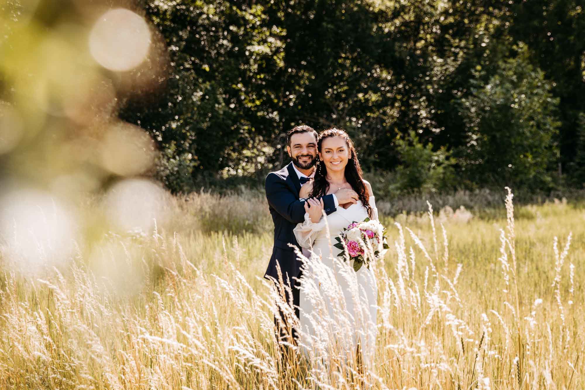 Das Brautpaar steht eng hintereinander mit Blick zum Fotografen. Links im Bild ist im Vordergrund ein Busch und im Hintergrund ist Wald und Feld zu sehen.