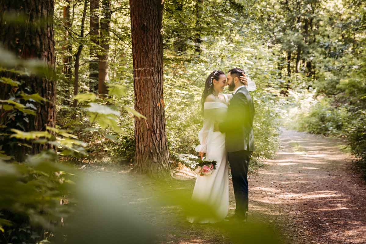 Das Brautpaar steht sich eng gegenüber. Die Braut hat die linke Hand am Kopf des Bräutigam platziert. Brautstrauß in der rechten Hand der Braut. Wald im Hintergrund.