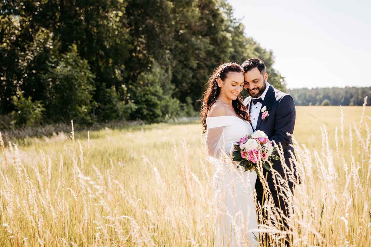 Das Brautpaar steht eng nebeneinander und hin der Mitte des Brautpaares halten beide den Brautstrauss auf den sie auch blicken. Im Hintergrund und Feld und Wald zu sehen.
