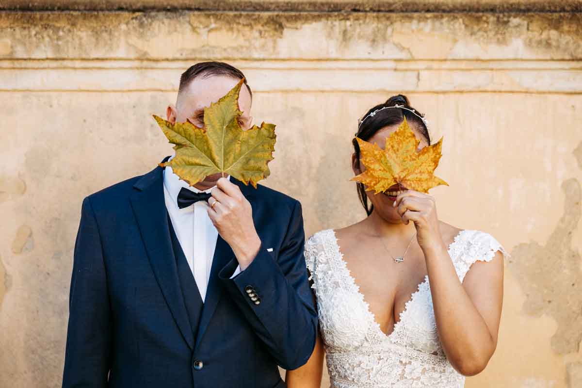 Das Brautpaar steht frontal zur Hauswand und haelt sich ein Kastanienblatt vor das Gesicht.
