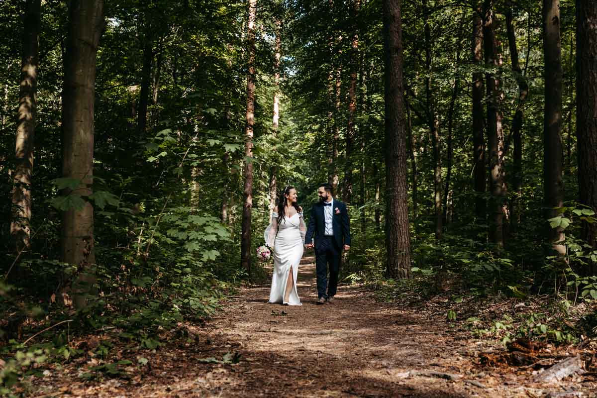 After Wedding Shooting mit dem Brautpaar im Wald. Sie kommen Hand in Hand auf die Fotografin zu und laecheln sich an.