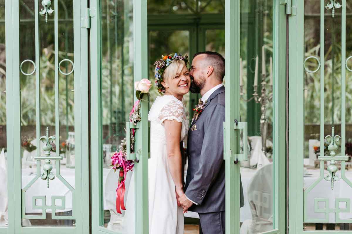Brautpaar steht in der geöffneten Tür vom Pavillon den Schlosses Diedersdorf. Der Bräutigam flüstert der Braut etwas ins Ohr. Sie lächelt.