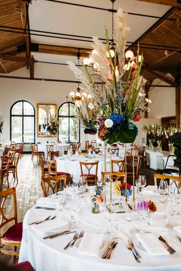 eingedeckte Tische für eine Hochzeit in der Location Cappuccino Gatow. Mächtiger Blumenschmuck in der Mitte der TIsche.