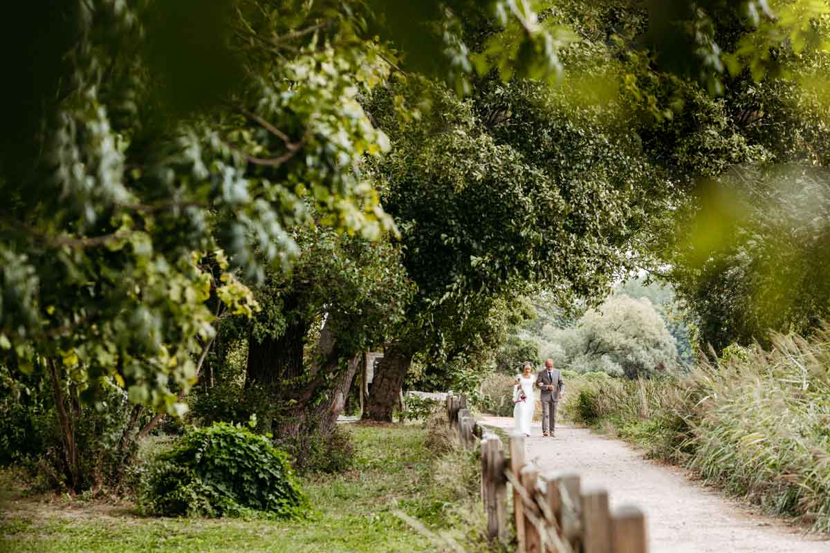 Der Brautvater begleitet die Braut zur freien Trauung zum Bräutigam. Links Bäume und rechts Büsche. Der Weg ist von einem Geländer getrennt.
