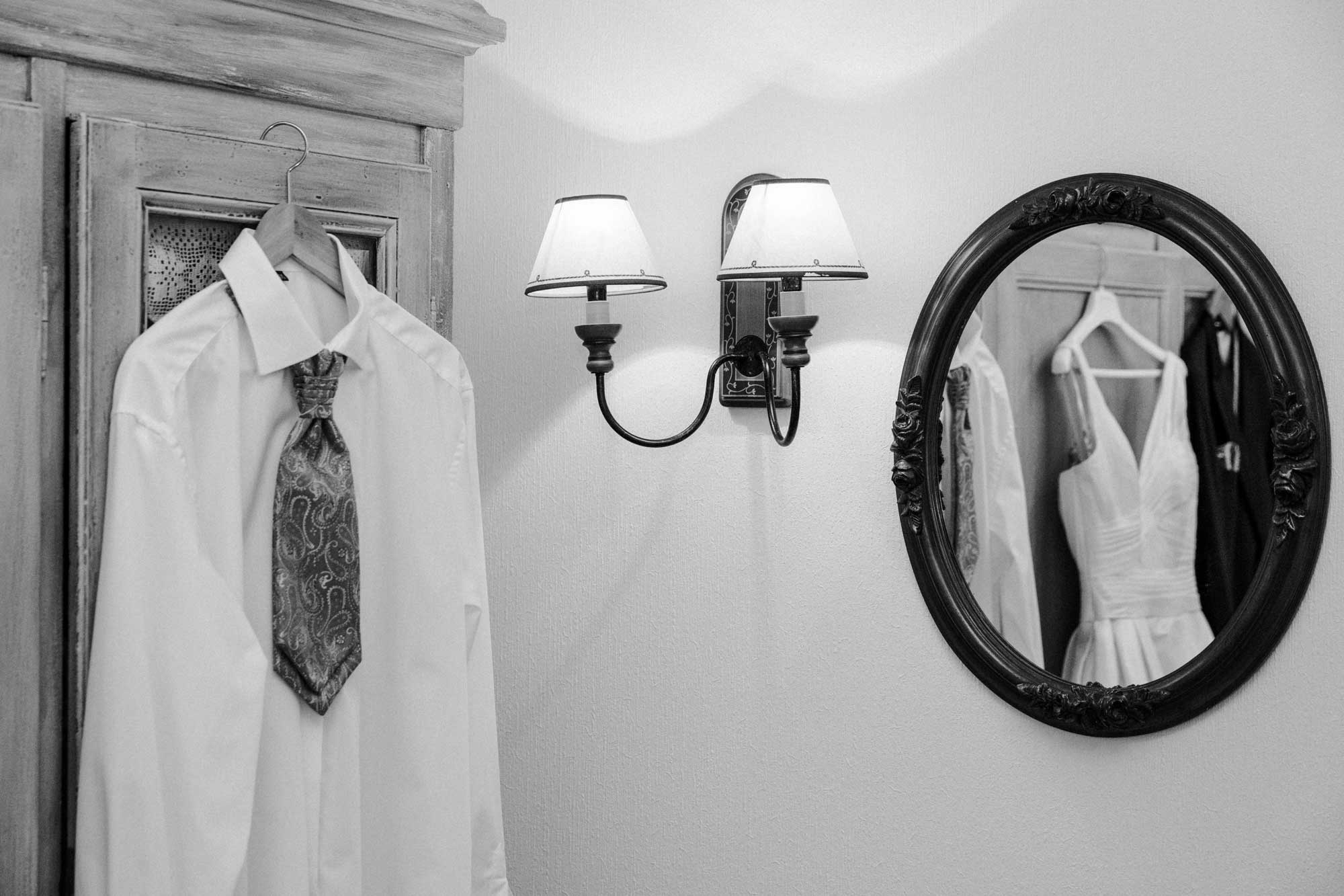 Das weiße Hemd mit Krawatte hängt auf einem Bügel am Schrank. Neben dem Schrank ist eine Wandleuchte und daneben ein runder Spiegel. In dem Spiegel spiegeln sich das Brautkleid und das Hemd mit Krawatte sowie der Anzug. Es ist eine schwarz weiß Aufnahme.