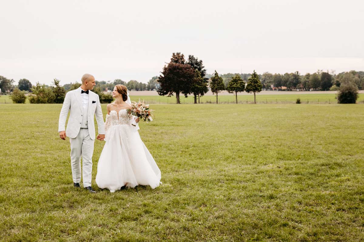Das Brautpaar läuft Hand in Hand über ein Feld. Sie lächeln sich an. Die Braut hält einen Brautstrauss in der rechten Hand.