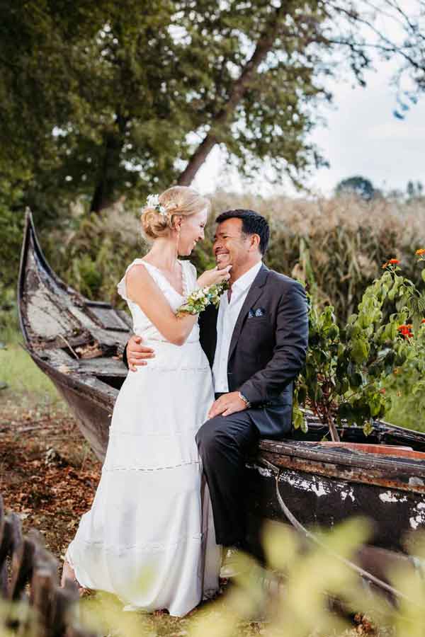 Der Bräutigam sitzt auf einer Gondel, die am Ufer geparkt ist. Die Braut sitzt auf seinem Schoss und sie lächeln sich an.