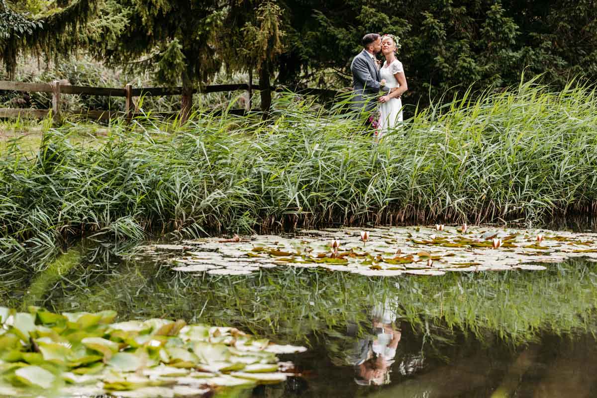 Brautpaar steht vor einem Teich, wo sie sich drin spiegeln und küssen sich. Gräser im Hintergrund.