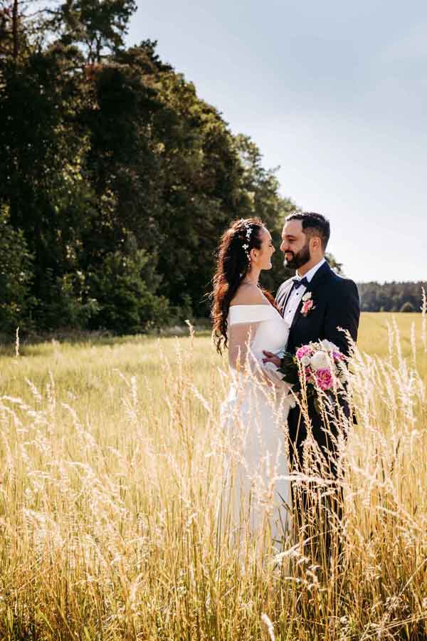 Das Brautpaar steht in einem Feld sich gegenüber. Sie lächeln sich an. Die Braut hält einen Brautstrauß in ihrer Hand.