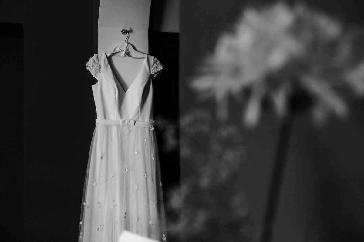 Brautkleid hängt auf dem Bügel an der Wand. Im rechten Vordergrund ist unscharf eine Blume. Schwarz-weiß Aufnahme.