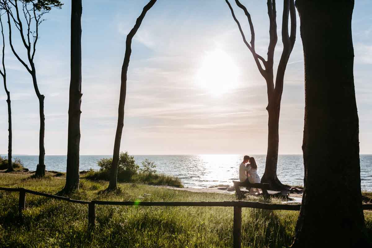 Das Paar sitzt bei dem Engagementshooting an der Ostsee auf einer Bank. Im Vordergrund sind Baeume und eine Wiese zu sehen und am Horizont geht die Sonne unter. Das Paar sitzt eng umschlungen und Kopf an Kopf.