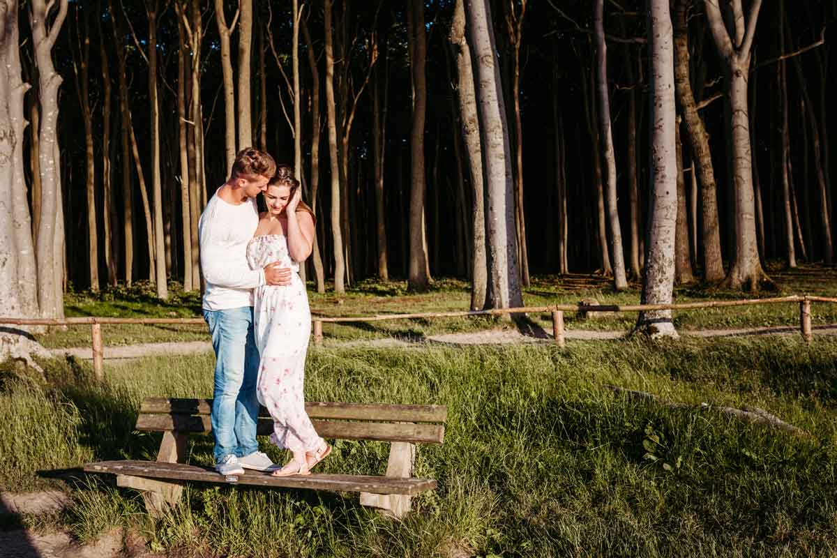 Das Paar steht auf einer Bank. Im Hintergrund ist Wiese und Wald. Es ist an der Ostsee aufgenommen. Man nennt die Gegend Maerchenwald. Das Paar steht sich eng gegenueber.