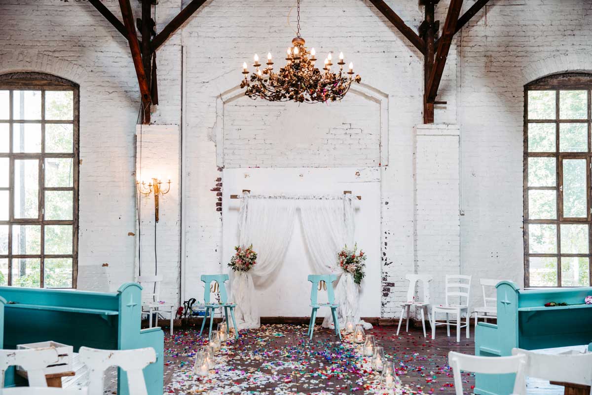 Traubogen mit Stühlen und Bänken für eine freie Trauung mit Blumenschmuck und Blüten auf dem Boden. Rechts und links Kerzen.